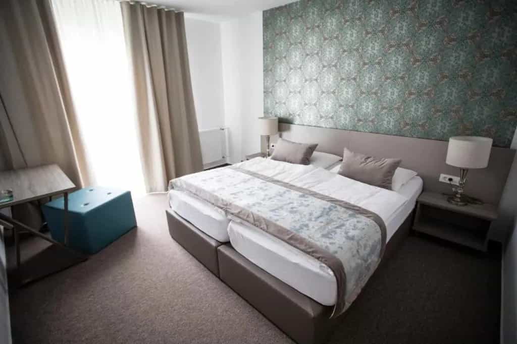 Hotel room with twin beds pushed together, patterned accent wall, and bright window with beige curtains.