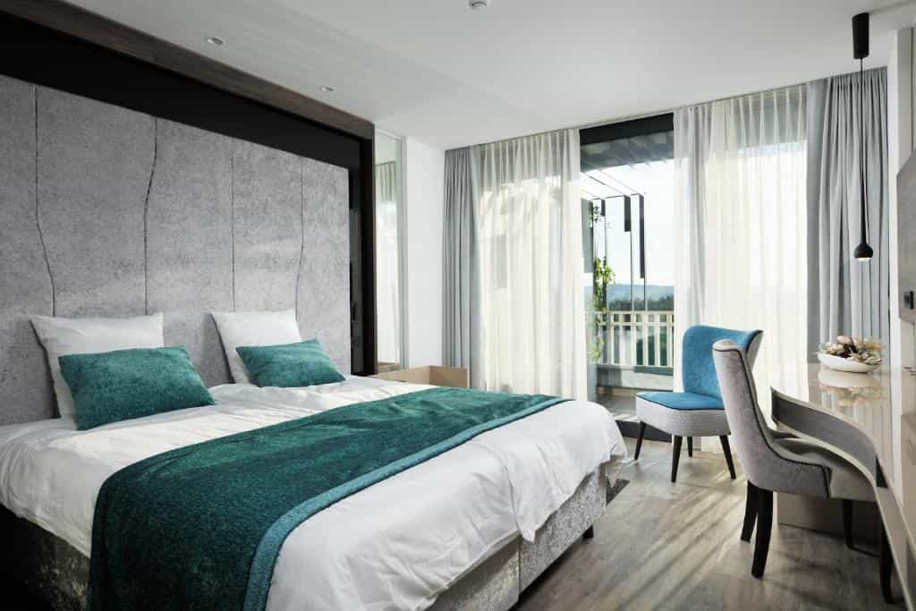 Modern hotel bedroom with white bedding, teal accents, and balcony view of green hills.