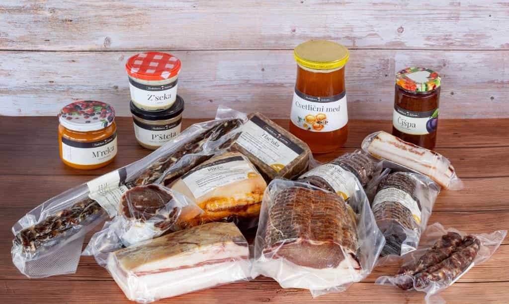 Assortment of vacuum-sealed cured meats, cheese, honey, and jams on a wooden surface.
