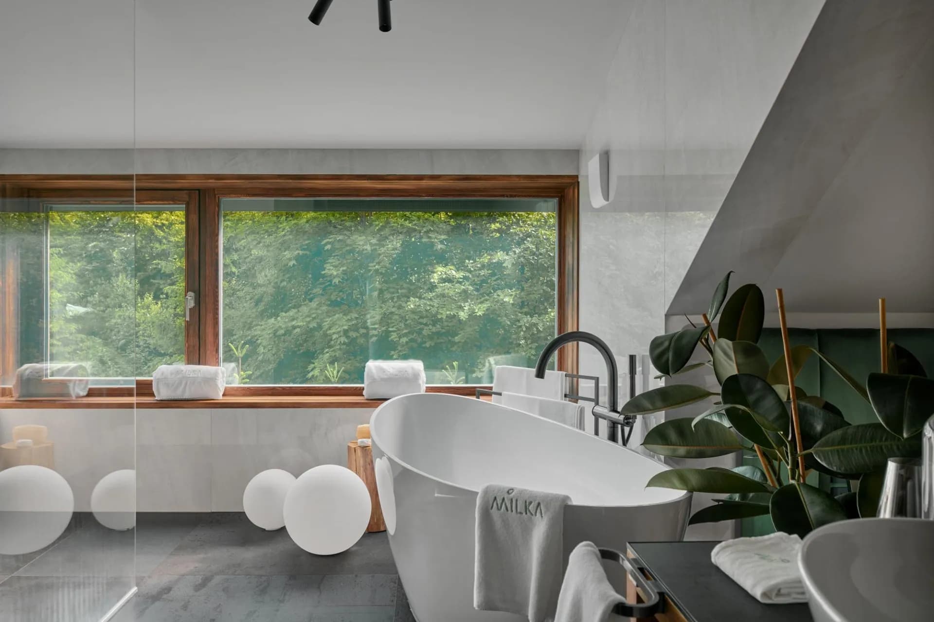 Modern bathroom with freestanding tub, large window overlooking dense green forest, and white sphere lights.