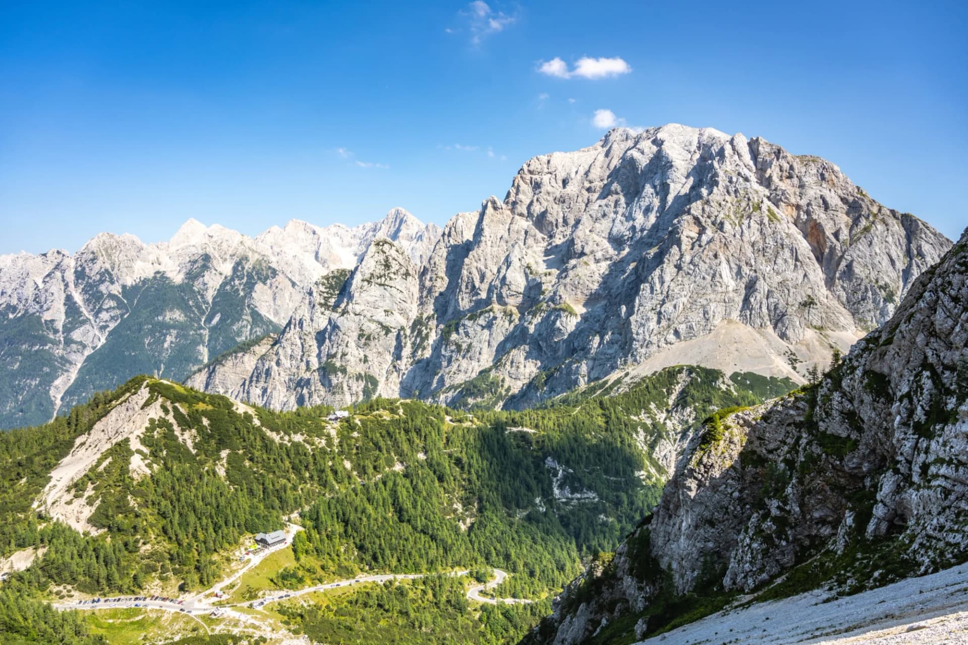 Rocky mountain peaks above green forested slopes with a winding road and building below.