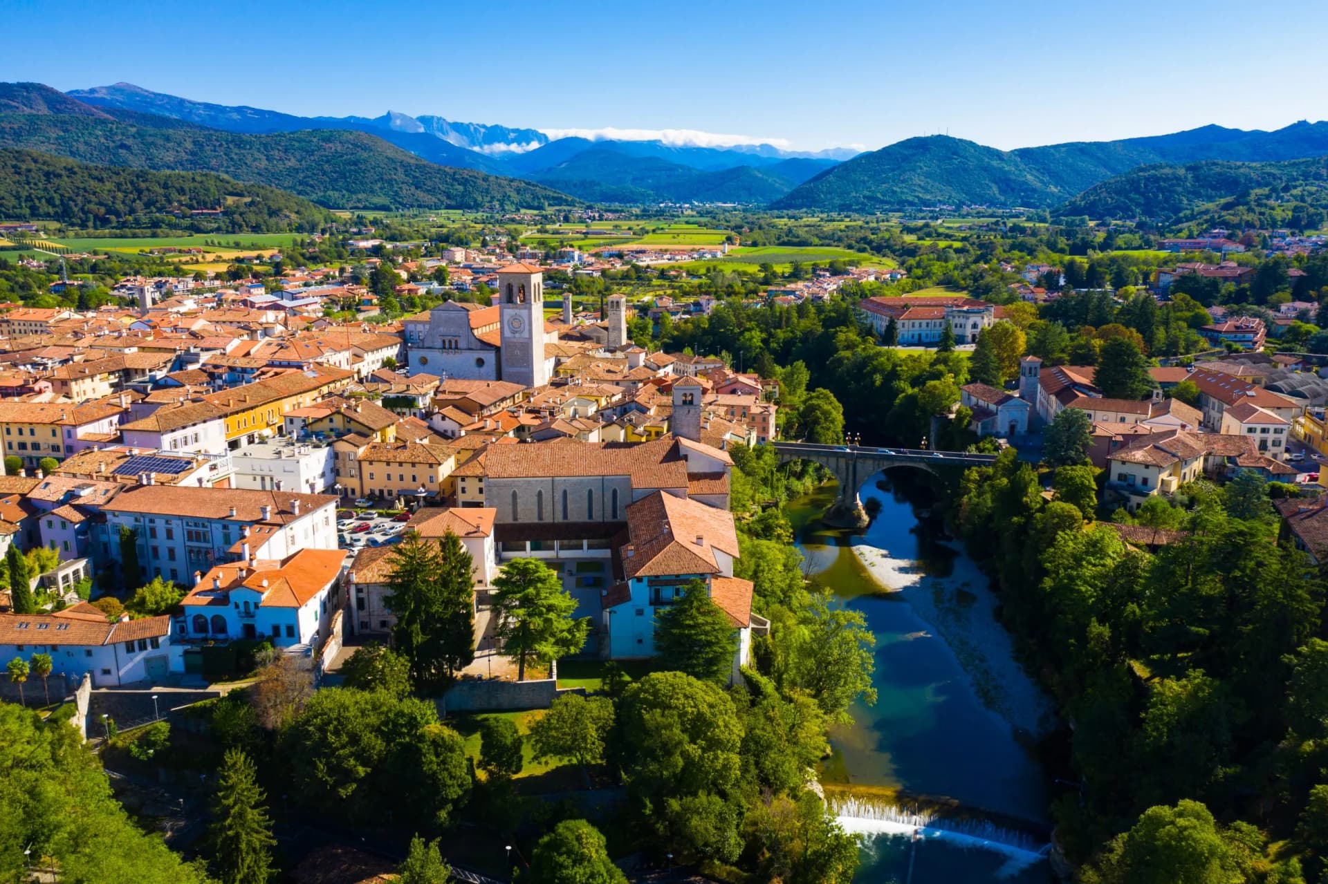 Aerial view of Cividale del Friuli town with terracotta roofs, river, and green mountains under blue sky.