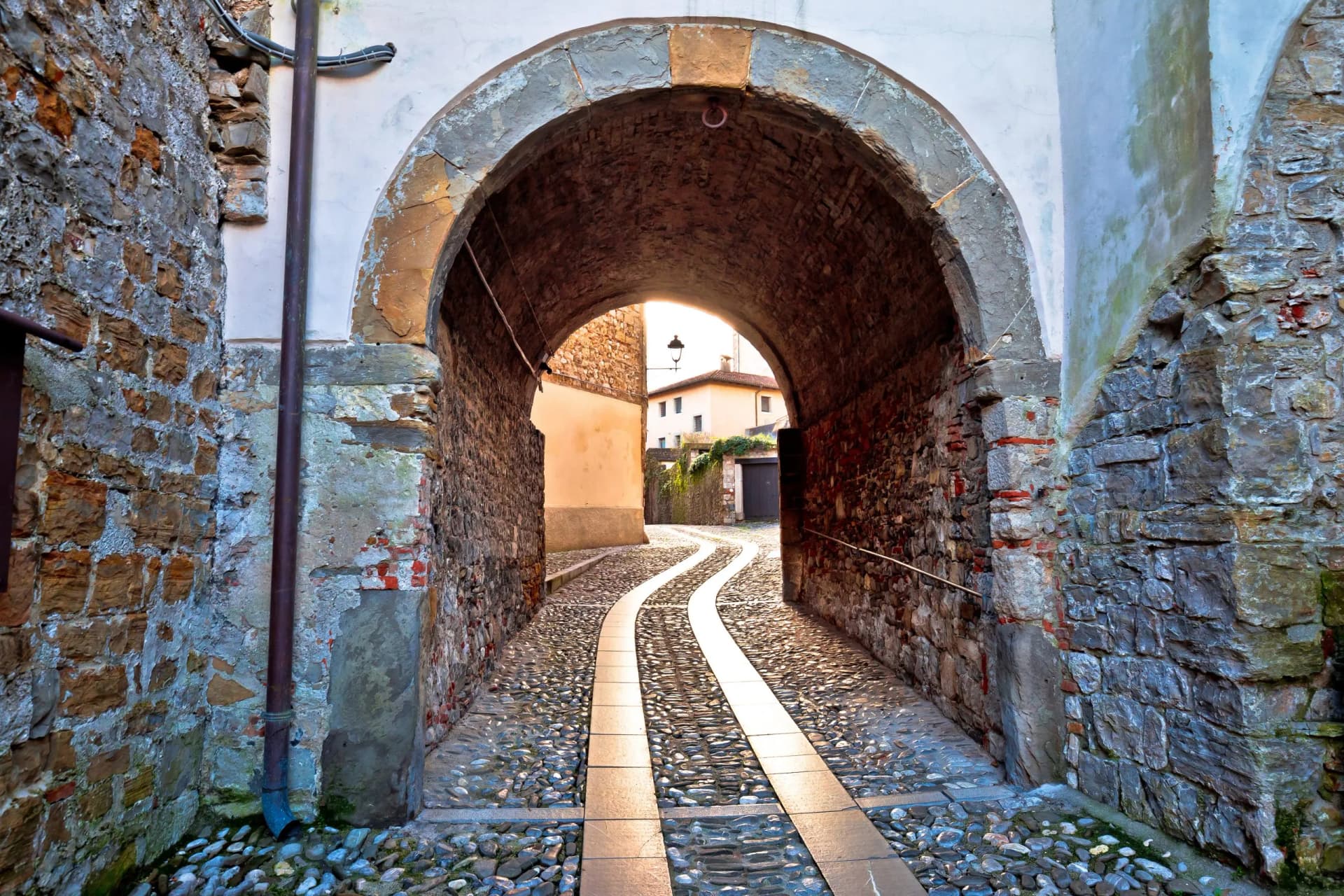 Cobbled alleyway under stone arch in Cividale del Friuli with sunlit winding path.