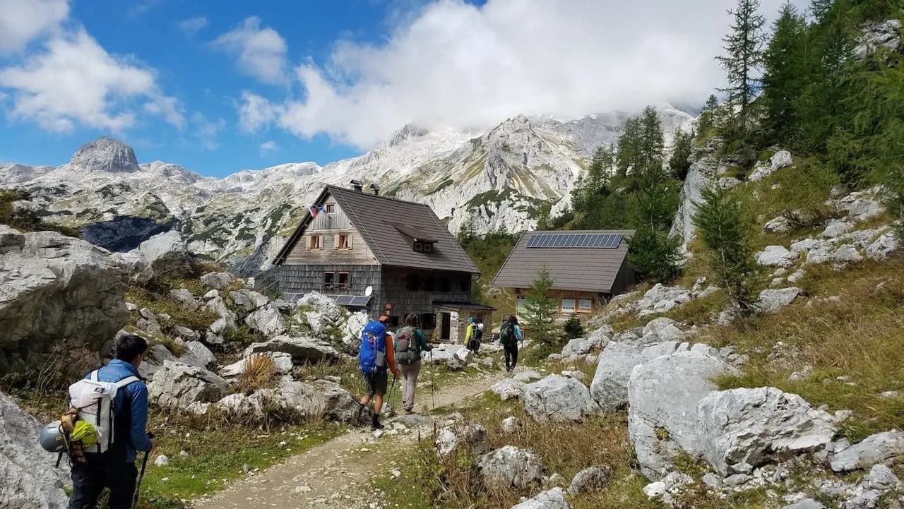 Hikers approach wooden mountain hut with solar panels below rocky peaks and blue sky