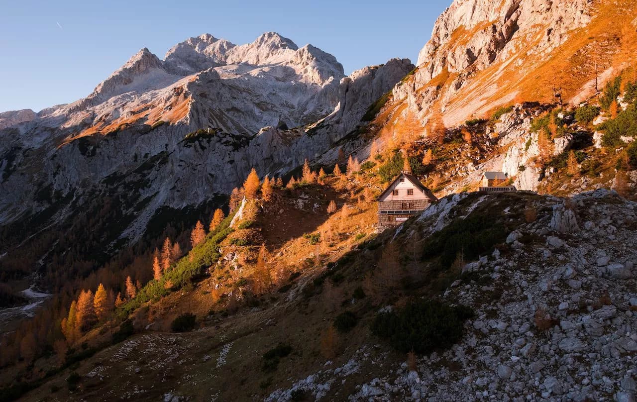 Vodnik Lodge nestled on a mountainside with Triglav peaks in the background during autumn.