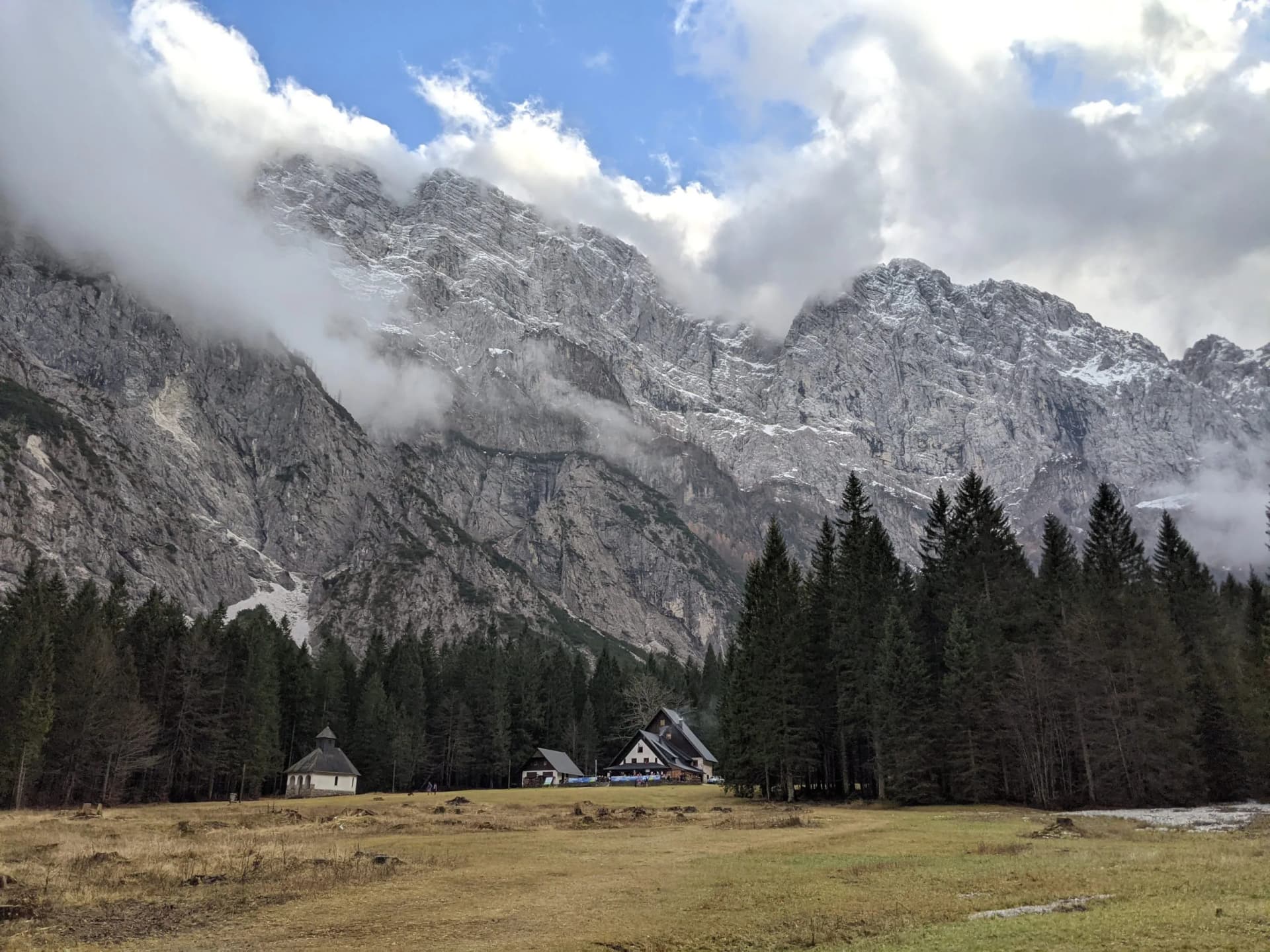 Alpine meadow with small buildings and chapel below rocky, cloud-covered mountains