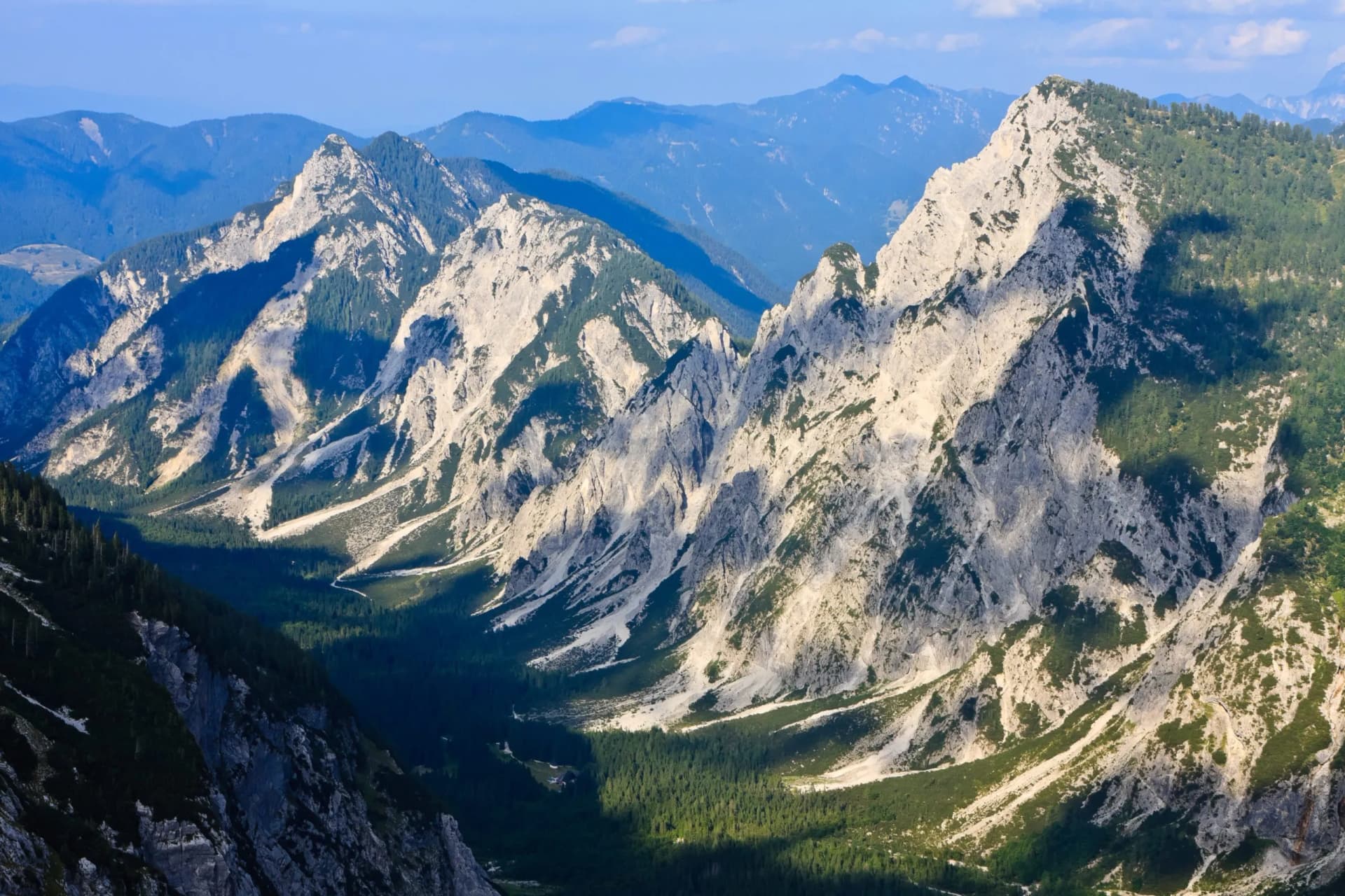 Rugged, sunlit limestone mountains with steep, forested slopes descending into a deep valley.