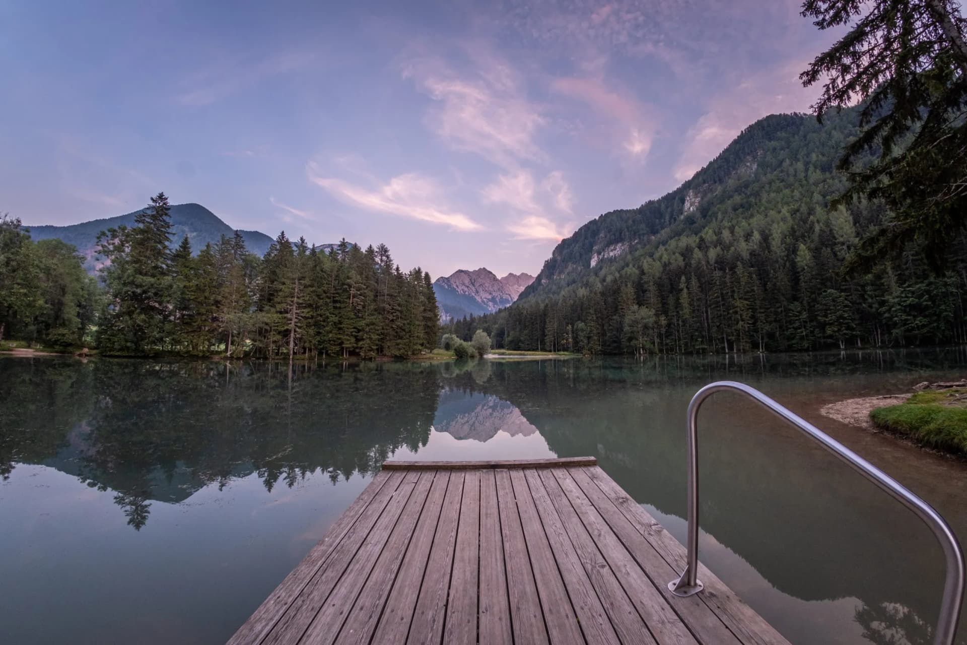 Wooden dock with metal railing on lake reflecting mountains and forest at dusk, Plansarsko Jezero