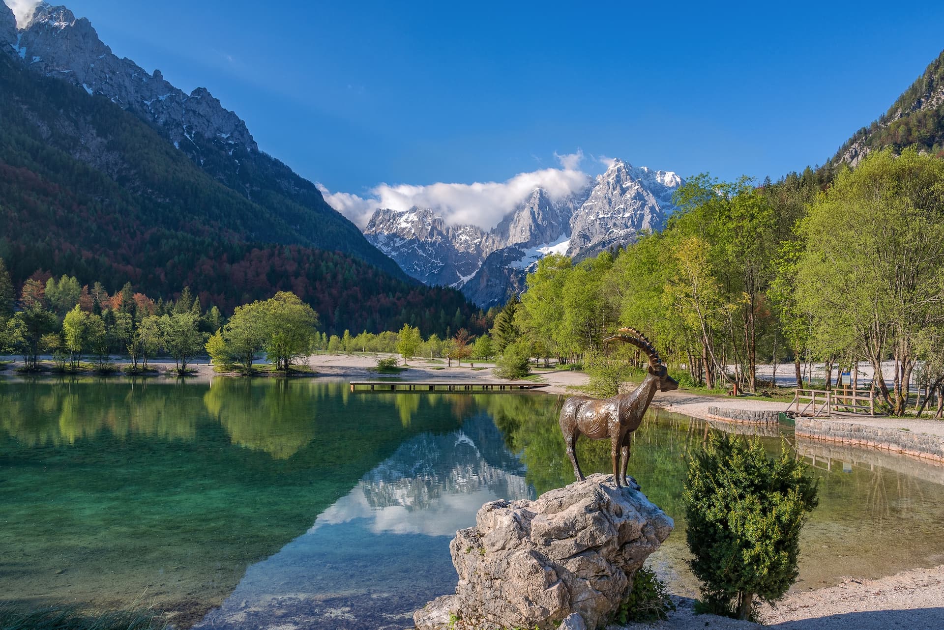 Bronze ibex statue by clear alpine lake with snow-capped mountains reflected in Kranjska Gora.