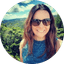 Woman in sunglasses smiling outdoors with lush green hills and blue sky background