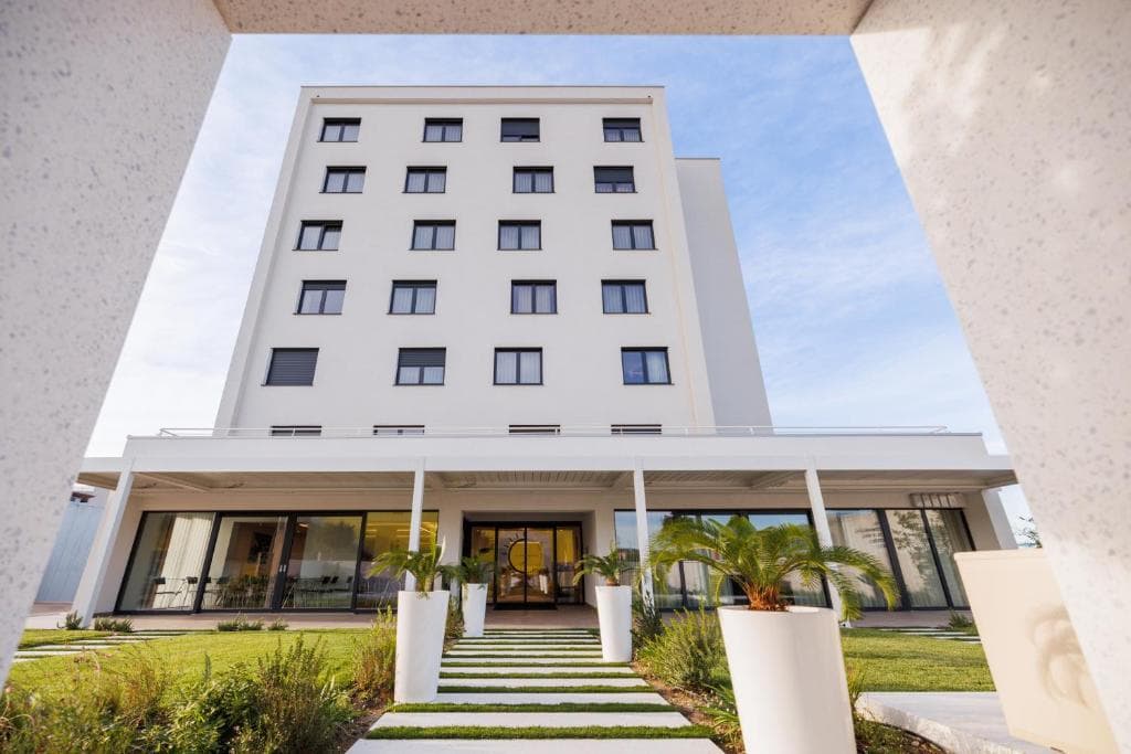 Modern white hotel building entrance framed by white architectural elements and green landscaping.
