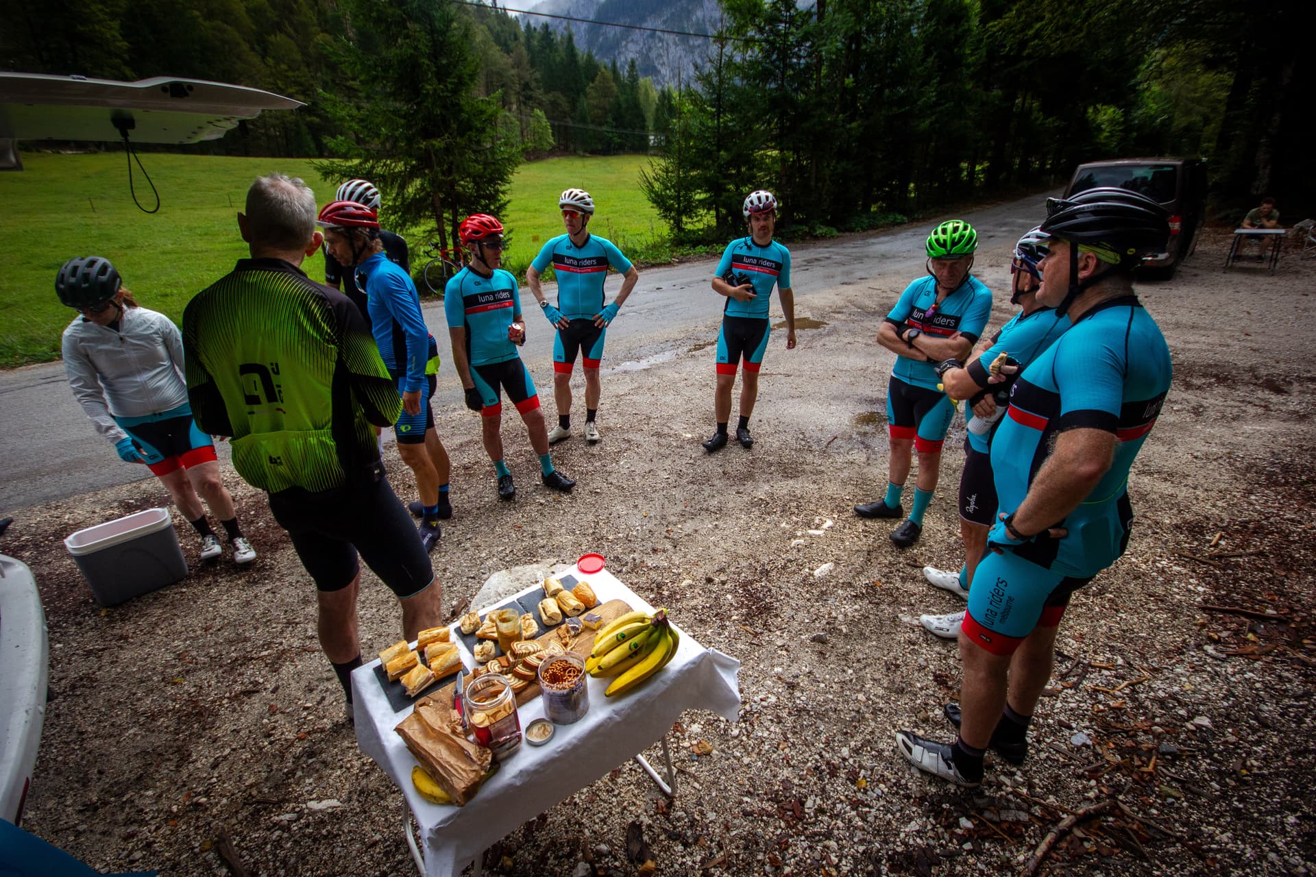 Cyclists taking a snack break with sandwiches and bananas near a forested mountain area in Slovenia.