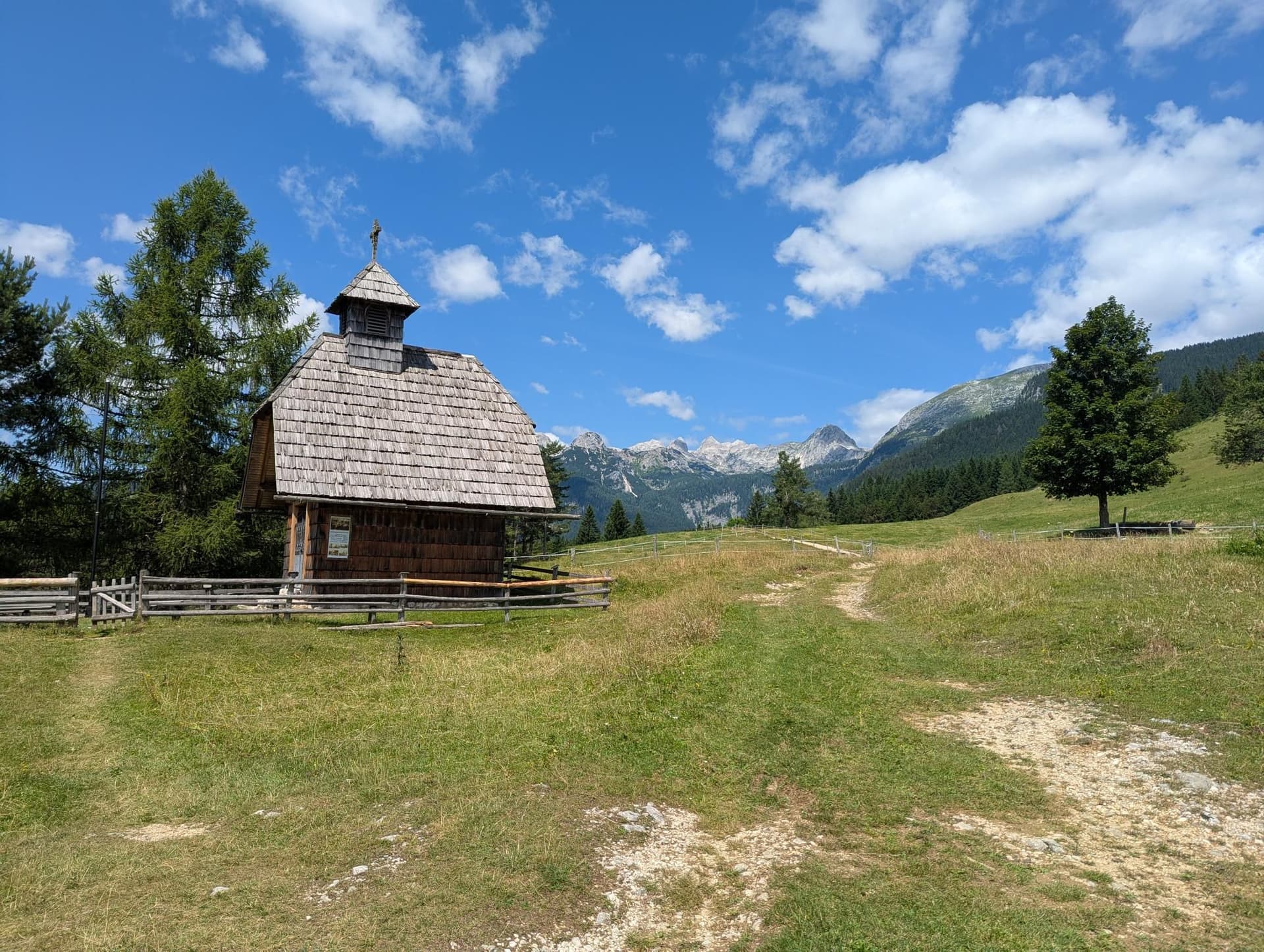 Small wooden chapel in a mountain meadow with Triglav peaks in the background.