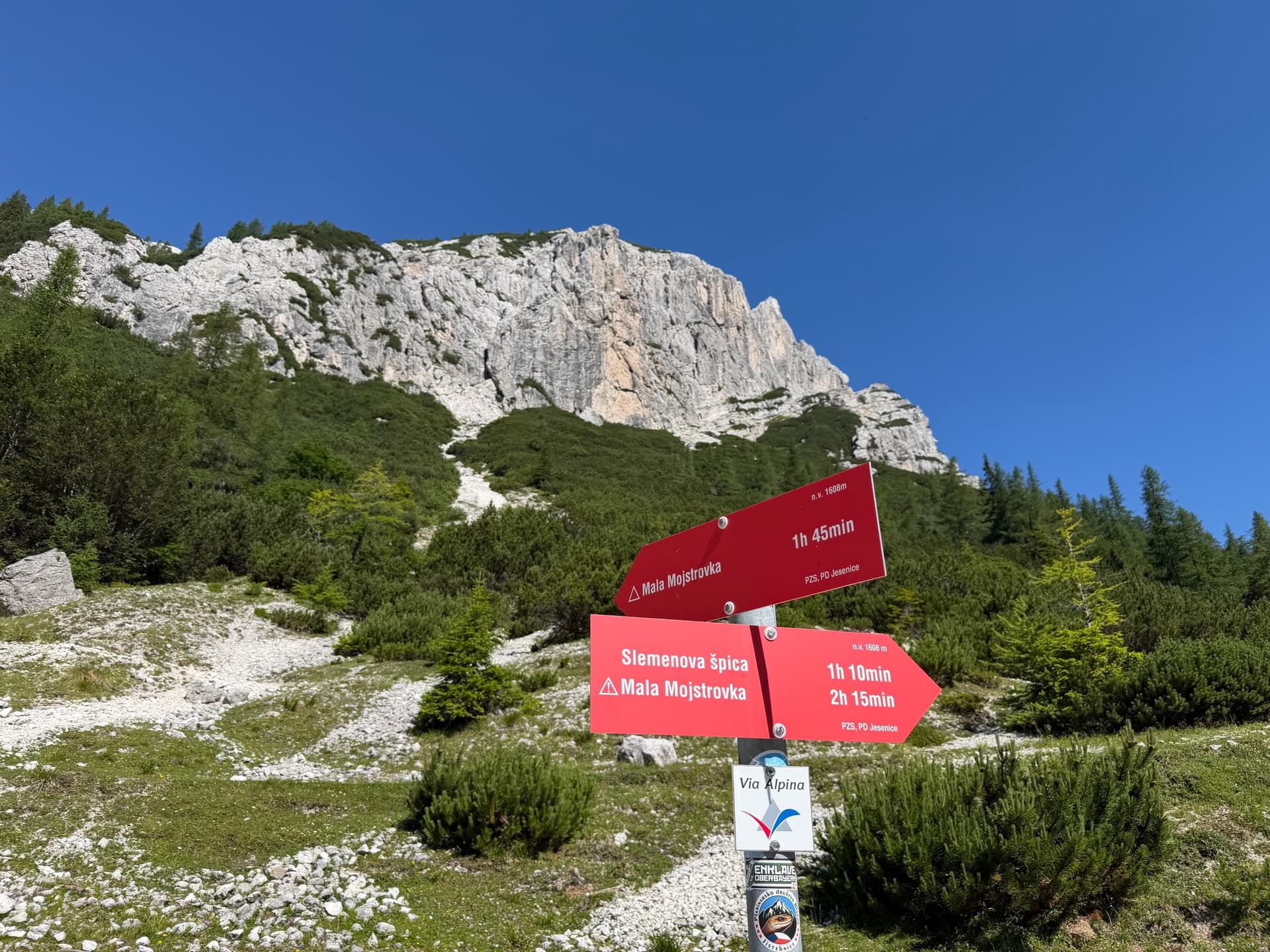 Red hiking signposts with trail destinations and walking times below limestone mountain peaks and blue sky