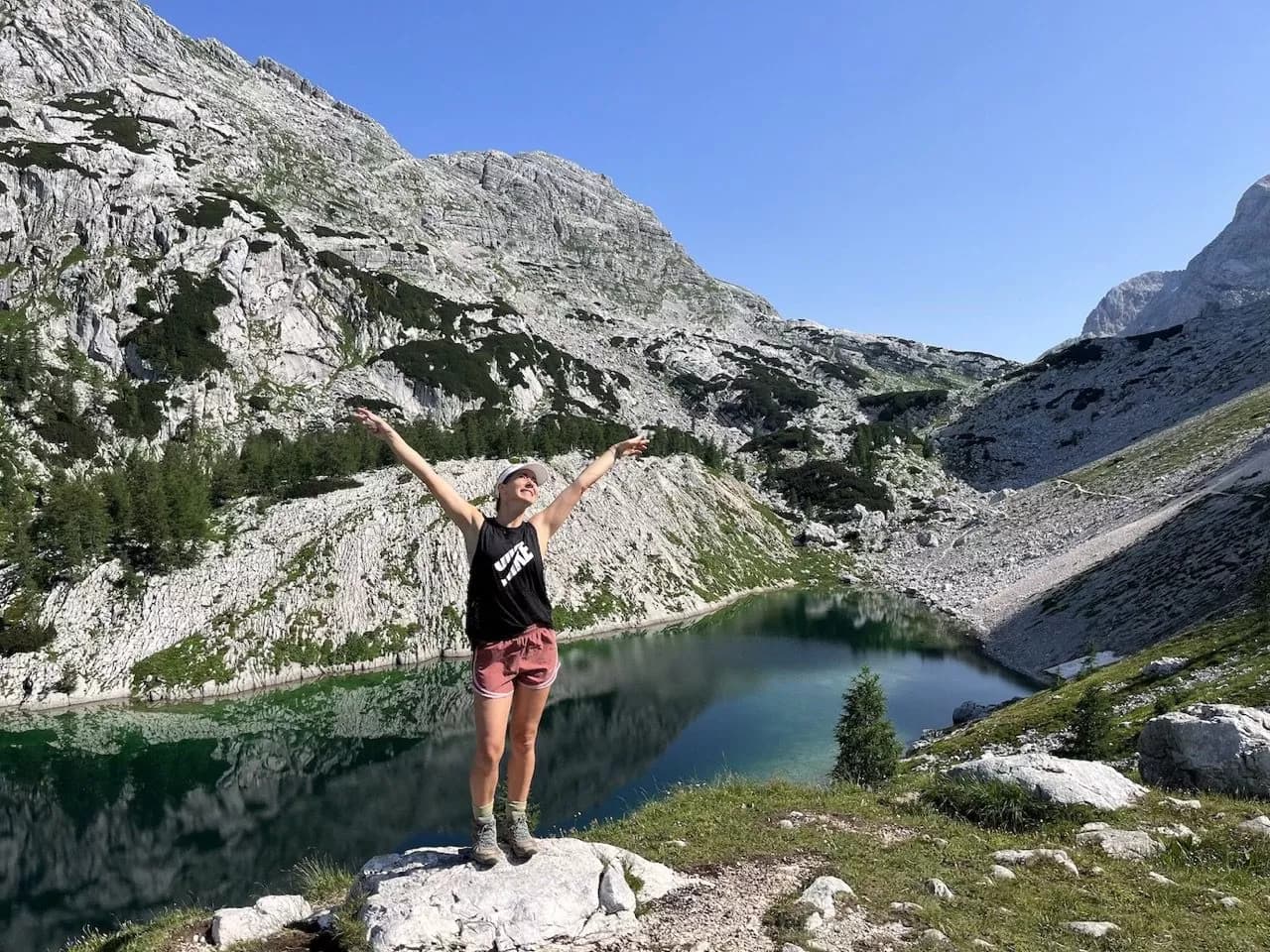 Hiker celebrating by alpine lake reflecting steep, rocky mountains under clear blue sky