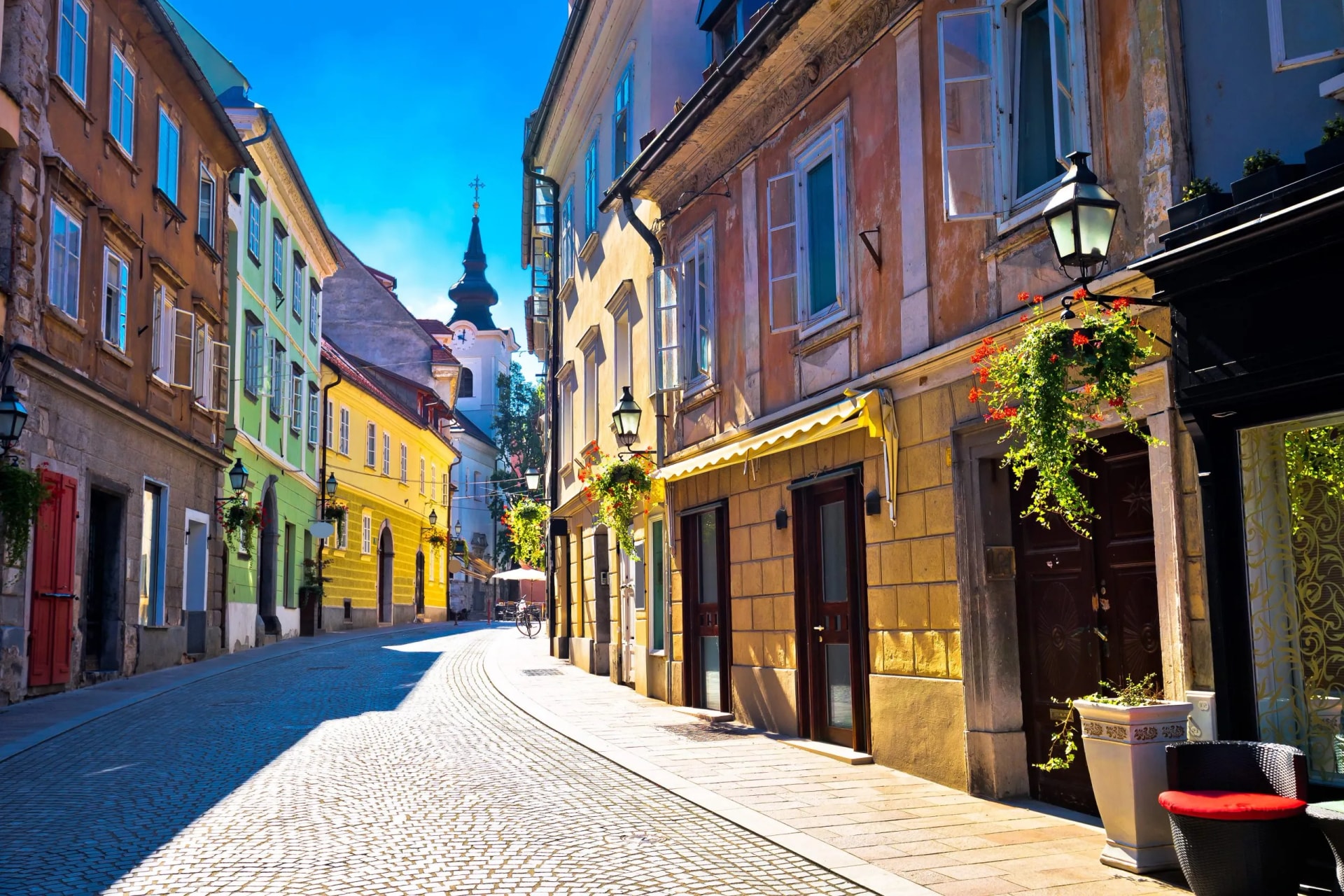 Cobblestone street in Ljubljana Old Town leading toward a church spire under a bright blue sky.