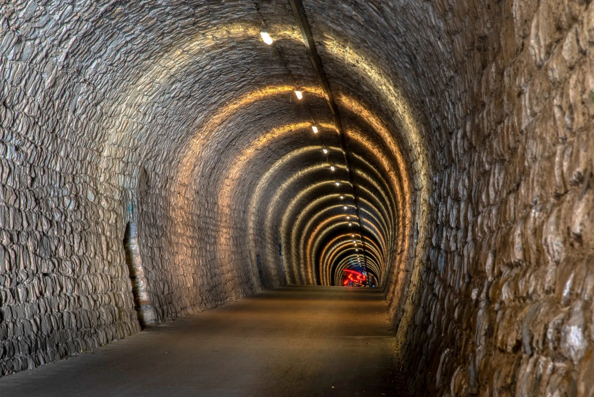 Stone-lined pedestrian tunnel with arched, illuminated ceiling leading toward red lights in the distance.