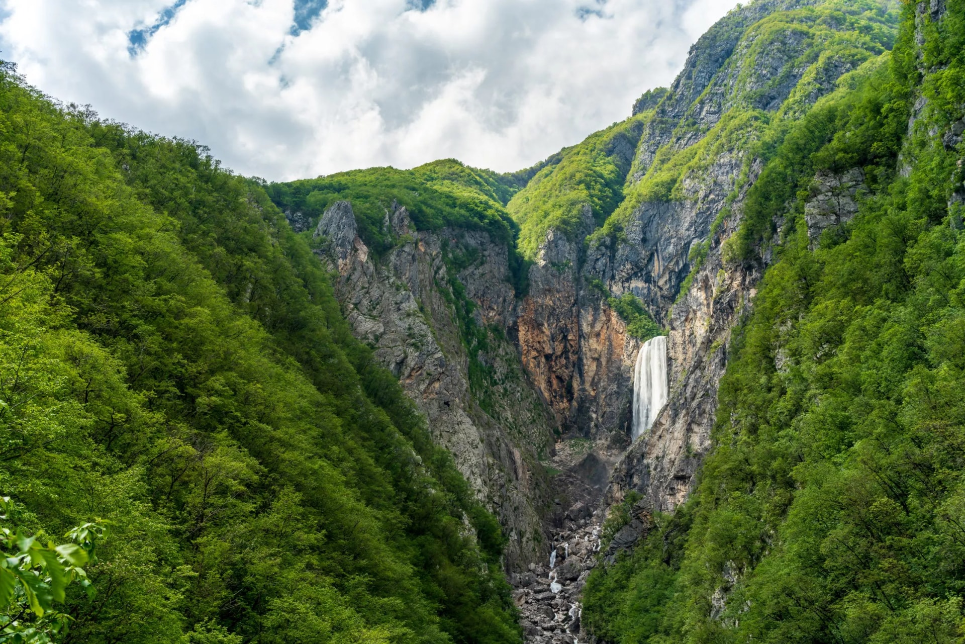 Slap Boka waterfall cascading down steep, green, forested cliffs under a cloudy sky in Slovenia.