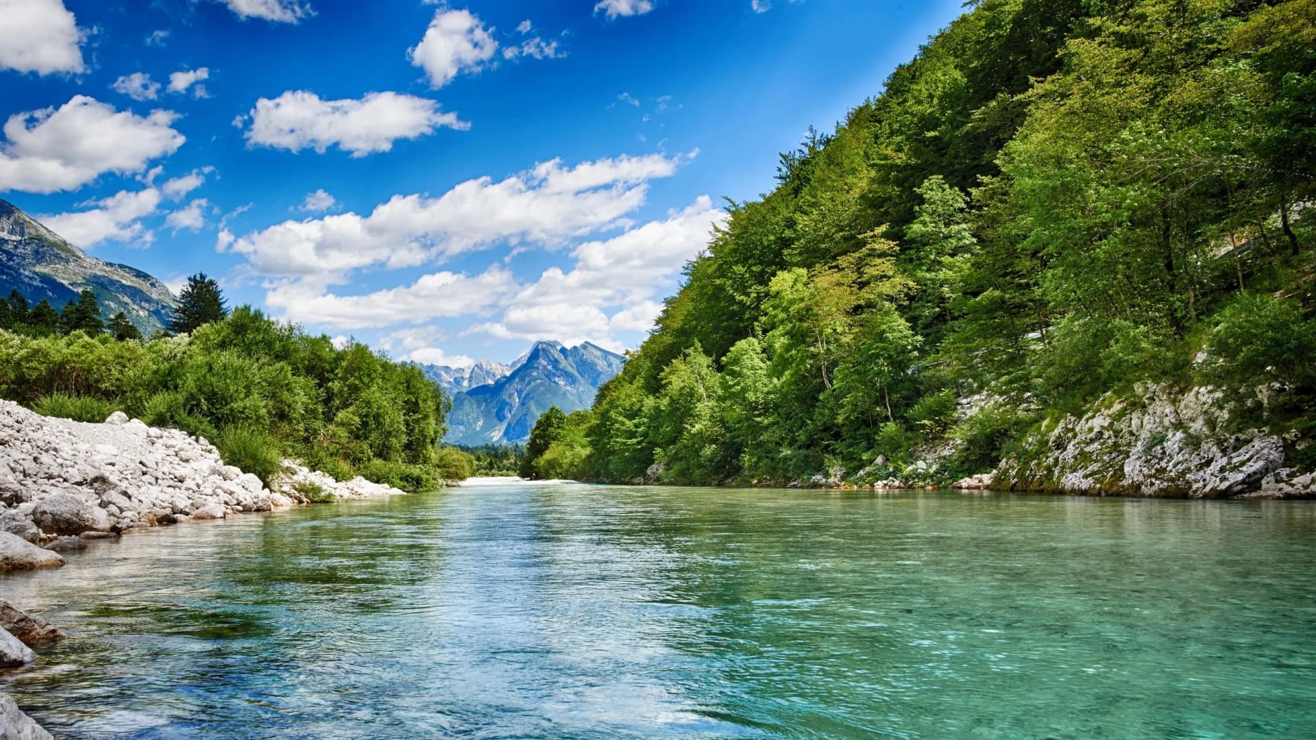 Clear turquoise river flowing between forested banks toward jagged, snow-capped mountains under a blue sky.