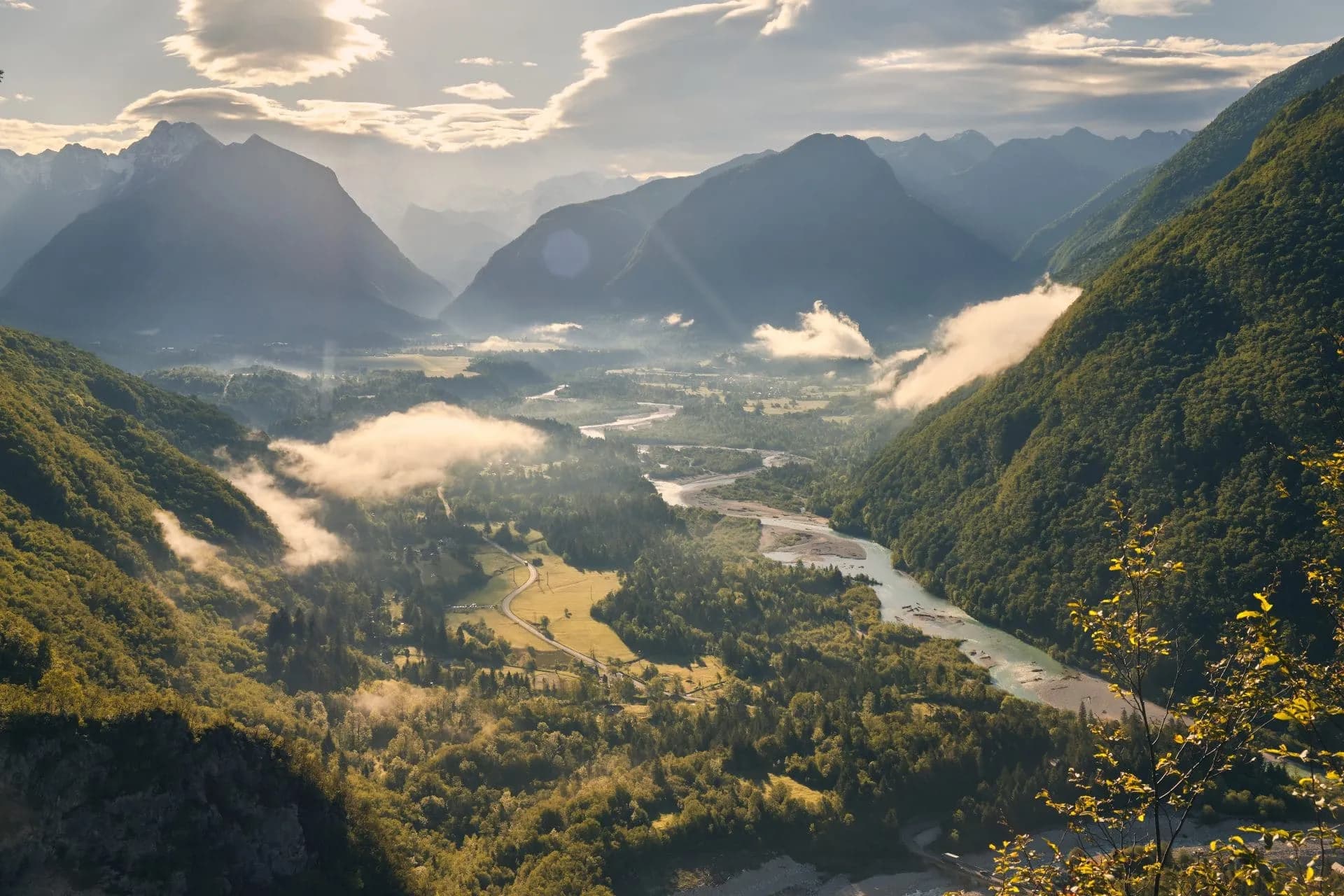 Panoramic view of Bovec area valley with winding river, green slopes, and morning mist among mountains.