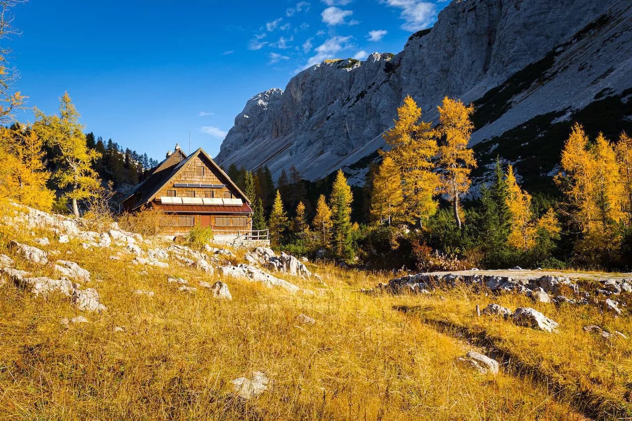 Triglav Lakes Lodge in autumn with yellow trees against a steep grey mountain under a blue sky.