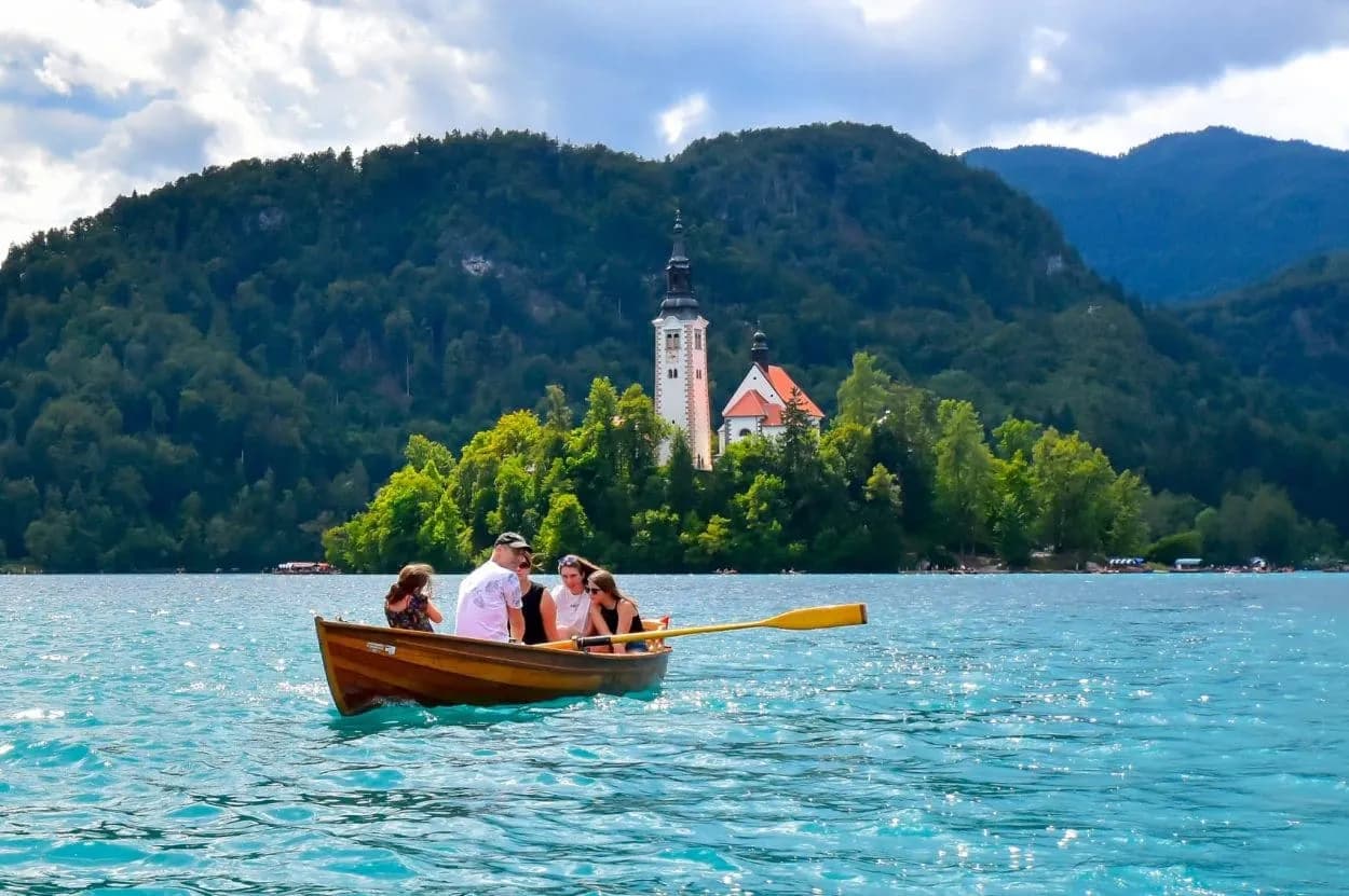 Rowing boat on turquoise water toward Bled Island church with forested mountains backdrop