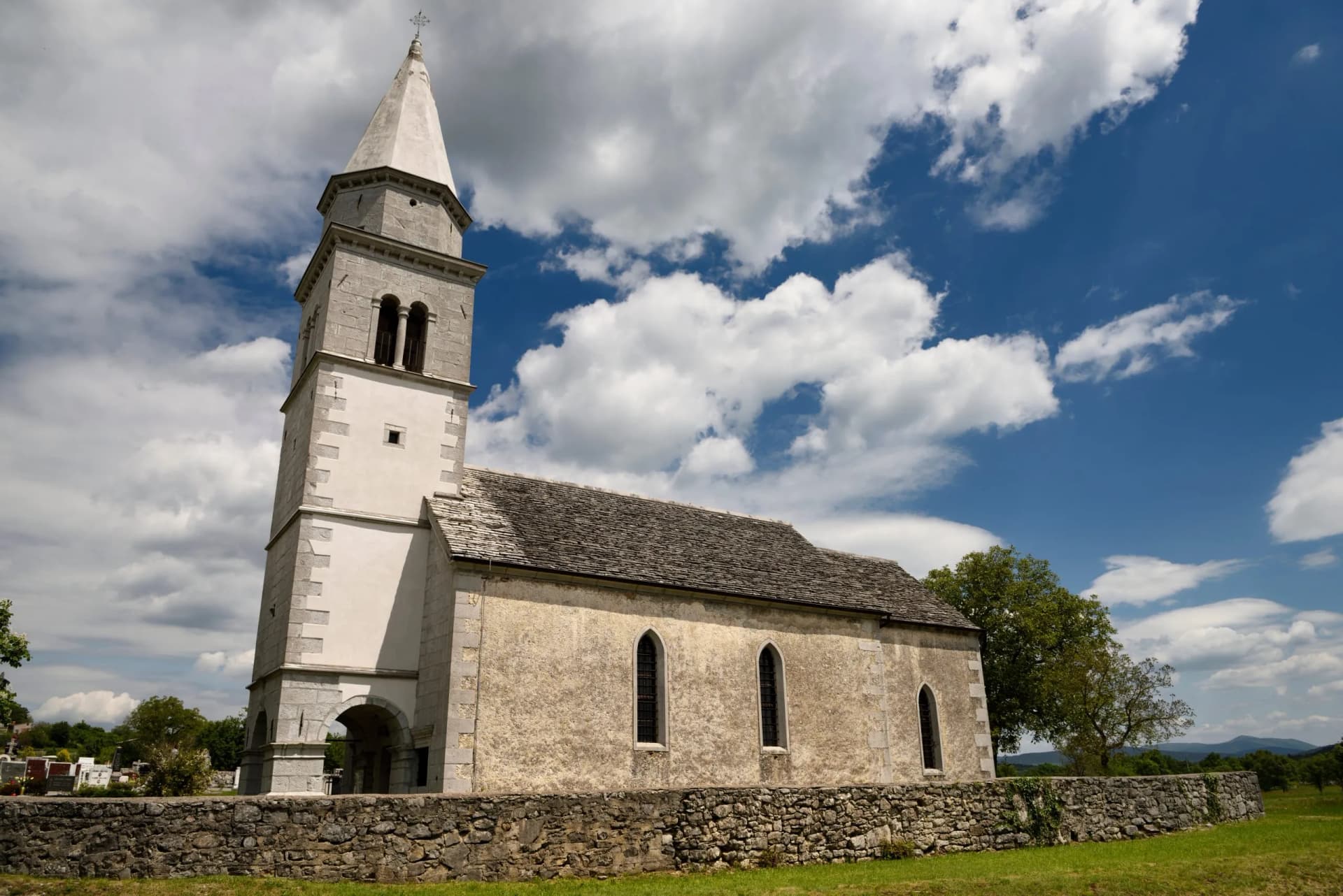 Church of the Holy Cross of Tomaj with stone bell tower under a dramatic blue sky with white clouds.