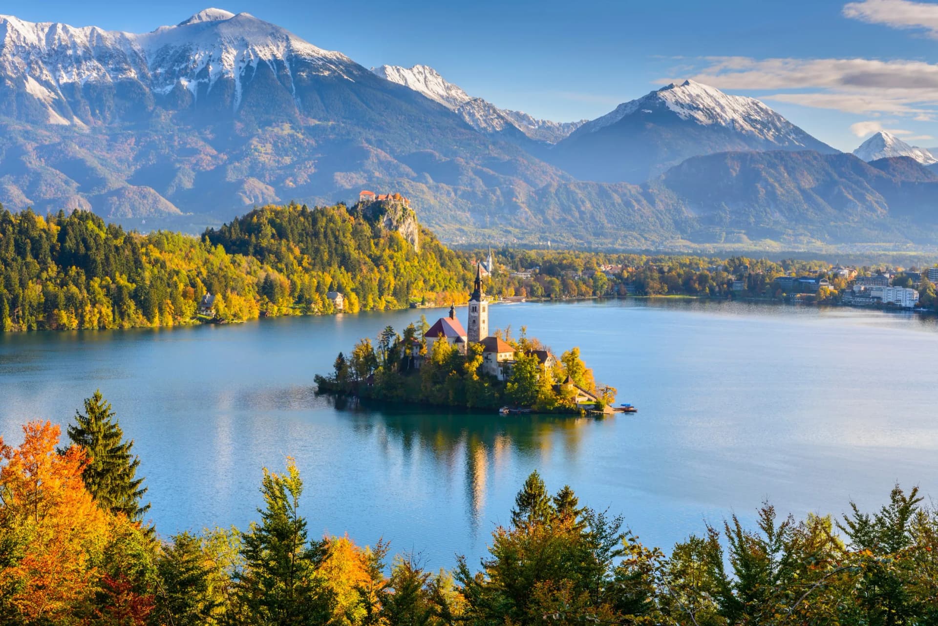 Panoramic view of Lake Bled island church with autumn trees and snow-capped mountains.