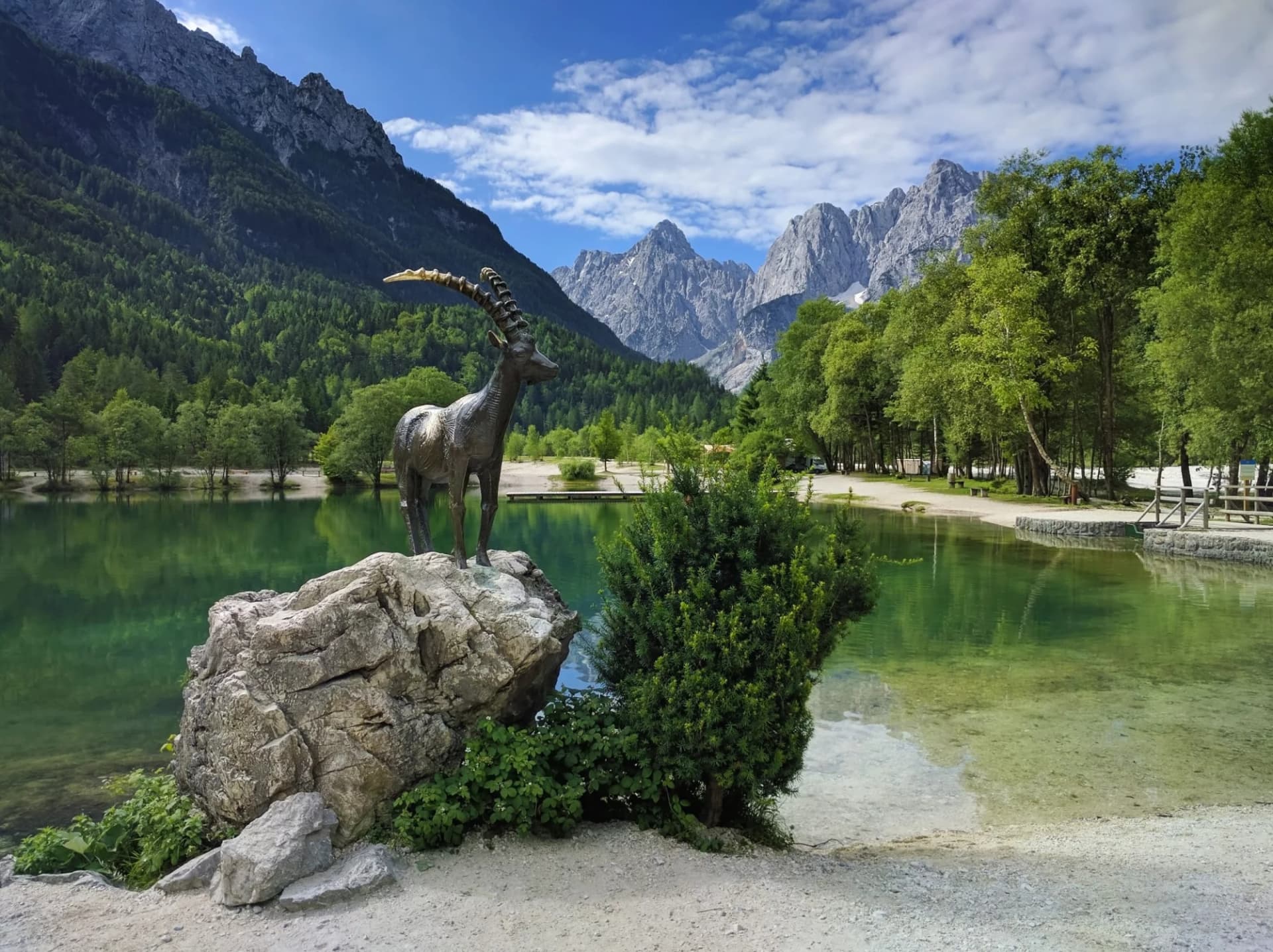 Mountain goat statue by Lake Jasna with green trees and Julian Alps in Kranjska Gora.