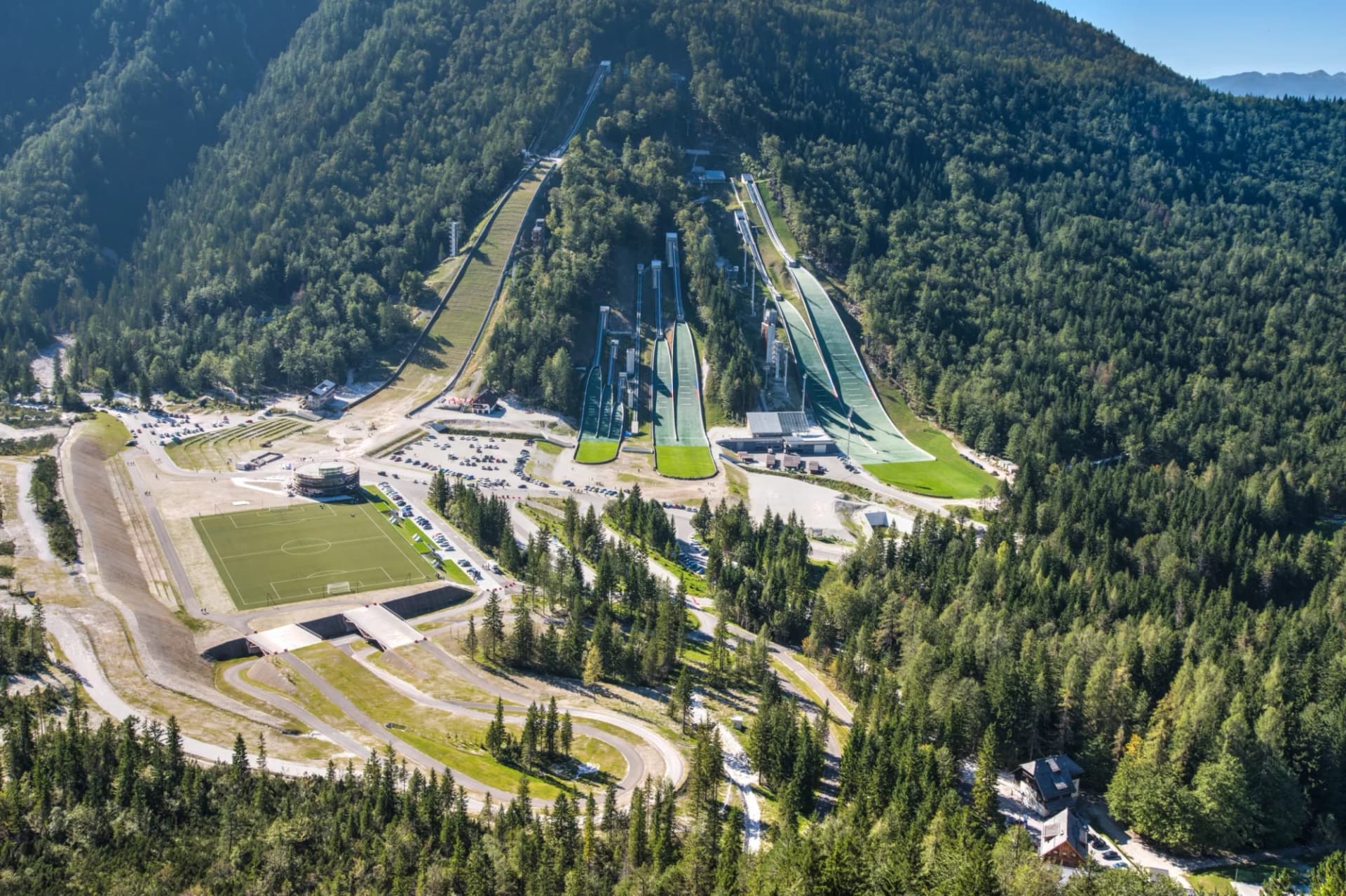 Ski jumping hills and soccer field nestled in a dense green forest mountain valley, Planica.