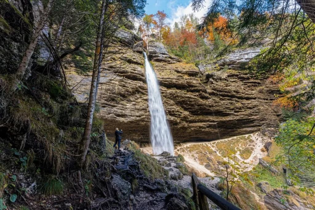 Peričnik waterfall cascading down layered rock face with autumn foliage and a hiker.