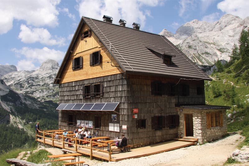 Vodnik's Hut (Vodnikov dom) with wooden exterior and terrace against rocky alpine mountains.