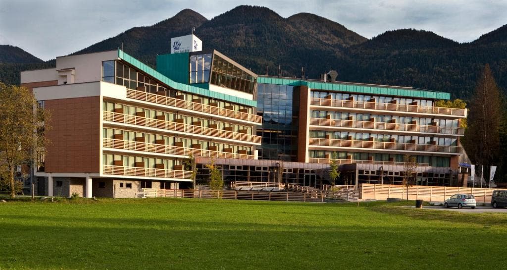 Bohinj Eco Hotel with balconies in front of forested mountains and a green lawn.