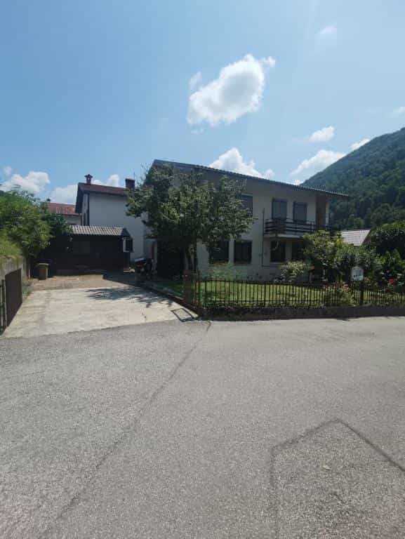 Two-story house with balcony and small yard, set against a steep, forested hill under a blue sky.