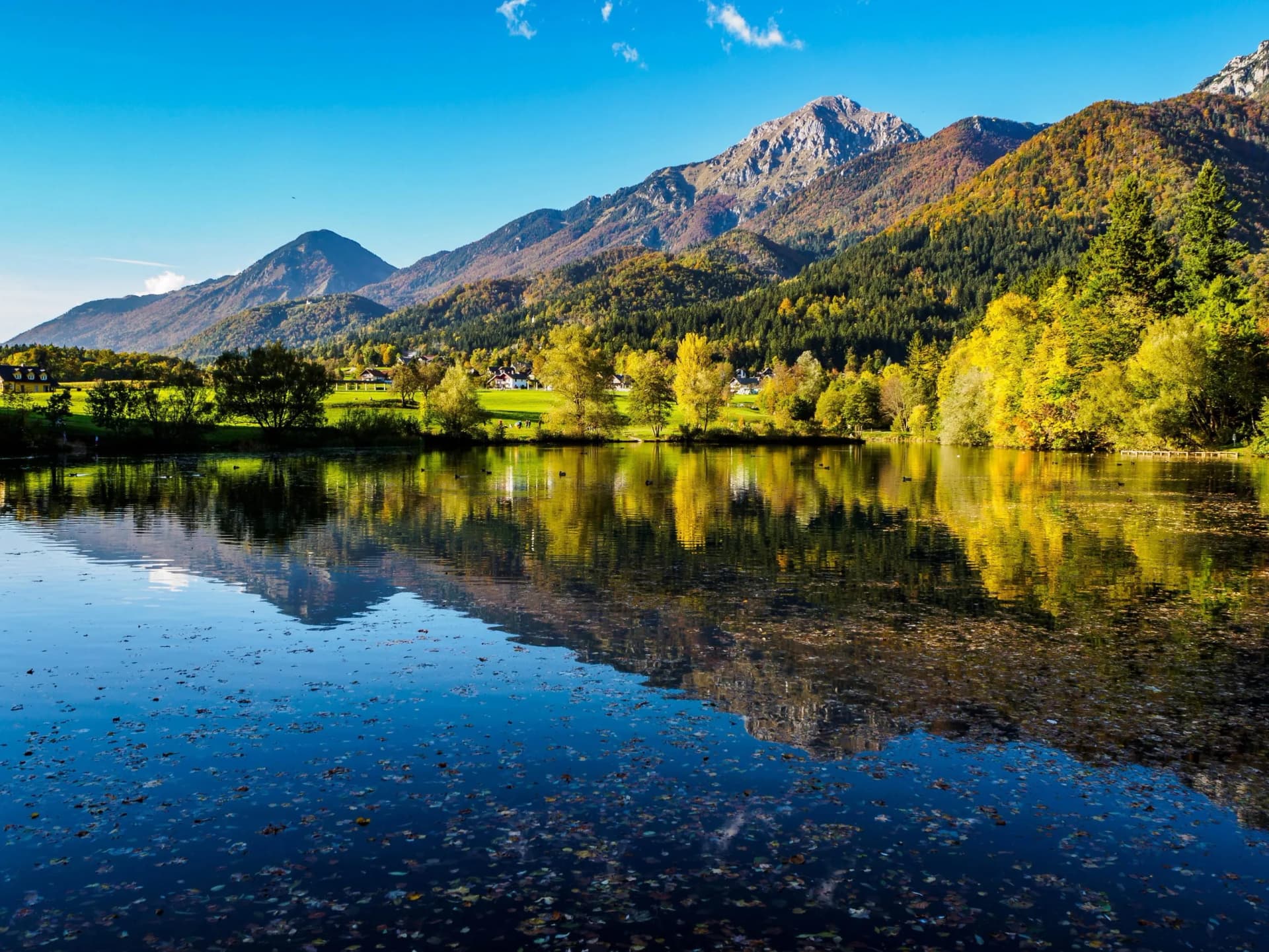 Alpine lake reflecting mountains with autumn foliage near small houses under a clear blue sky.
