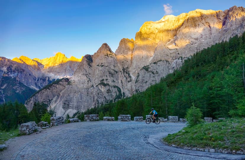 Active female tourist pedals her mountain bike up a cobblestone road at sunset.