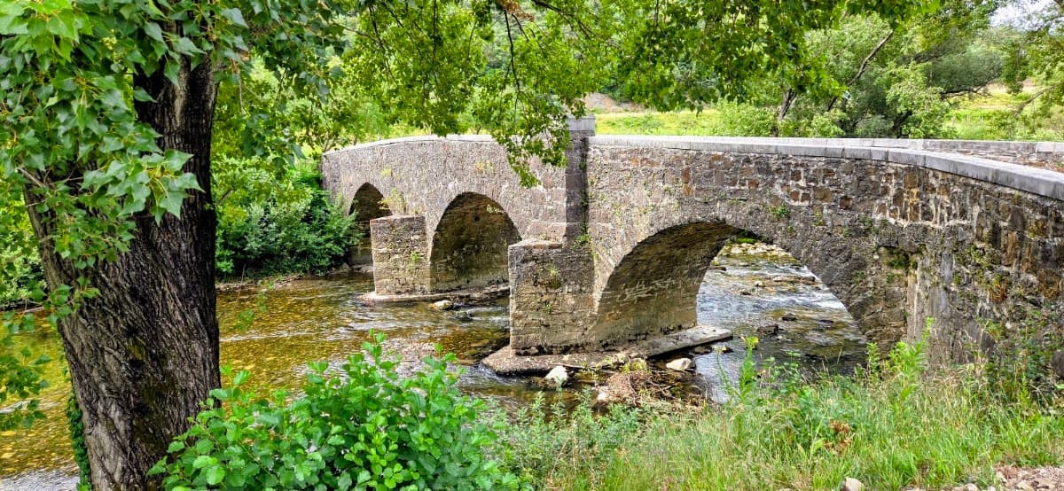Stone arch bridge over a shallow river with green trees and foliage