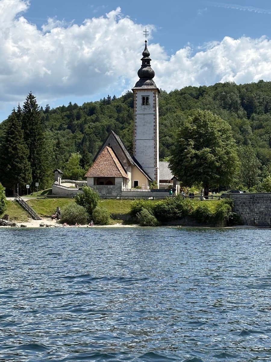 Historic stone church with tall bell tower on lake shore backed by forested mountains and blue sky