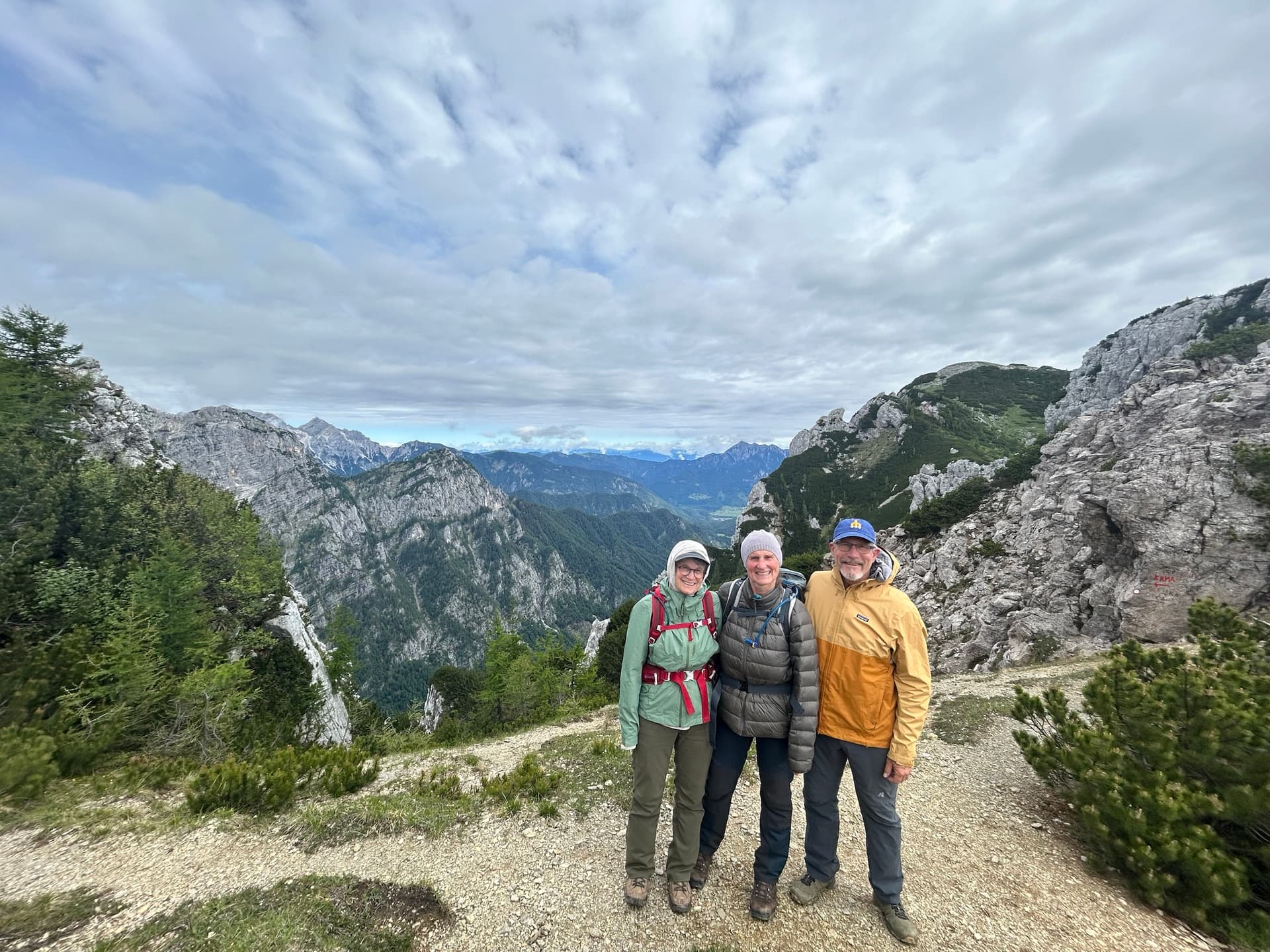 Three hikers standing on a mountain trail with rocky peaks and cloudy sky background