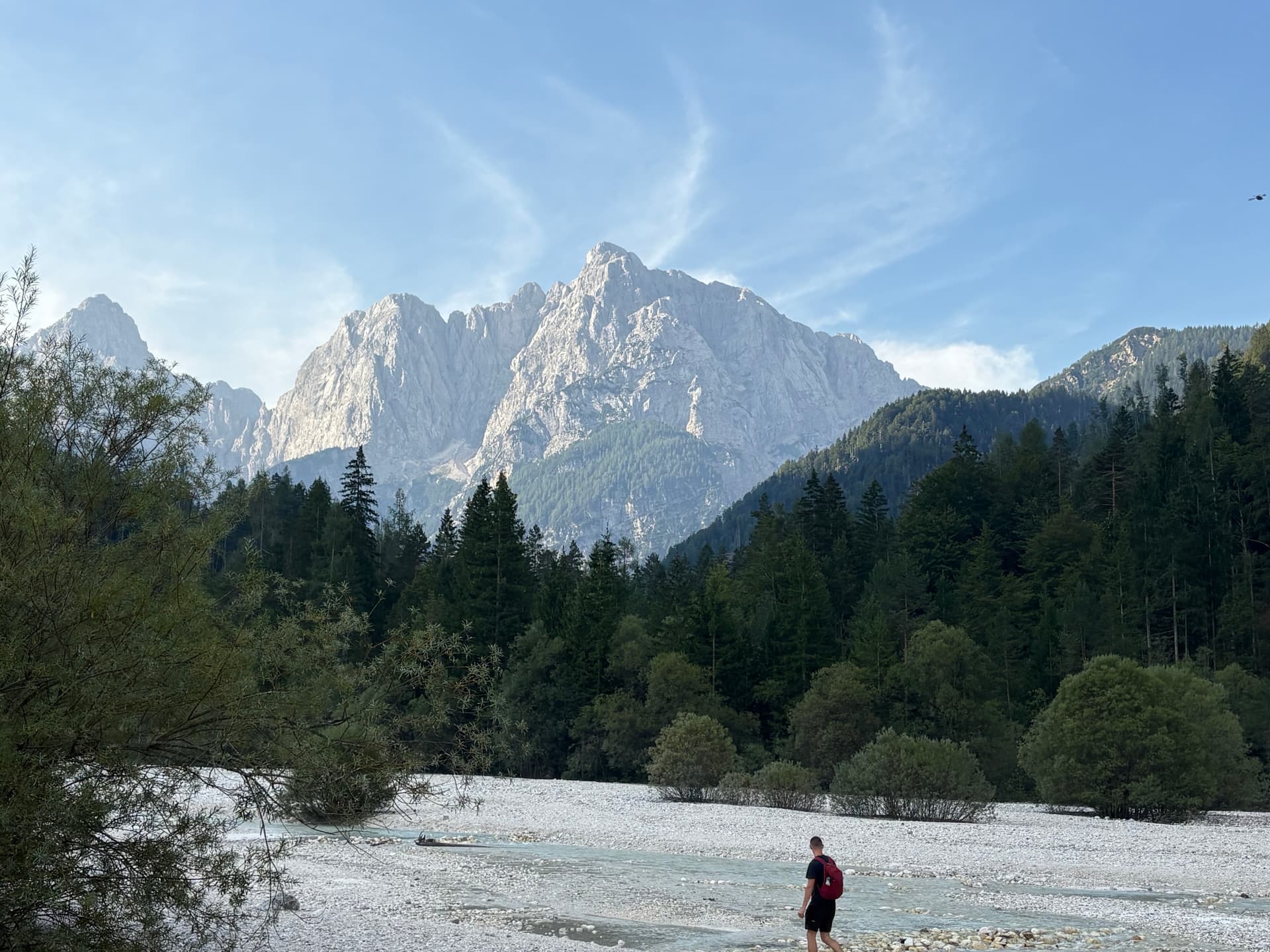Hiker walking on gravel riverbed below towering mountain peaks and pine forest