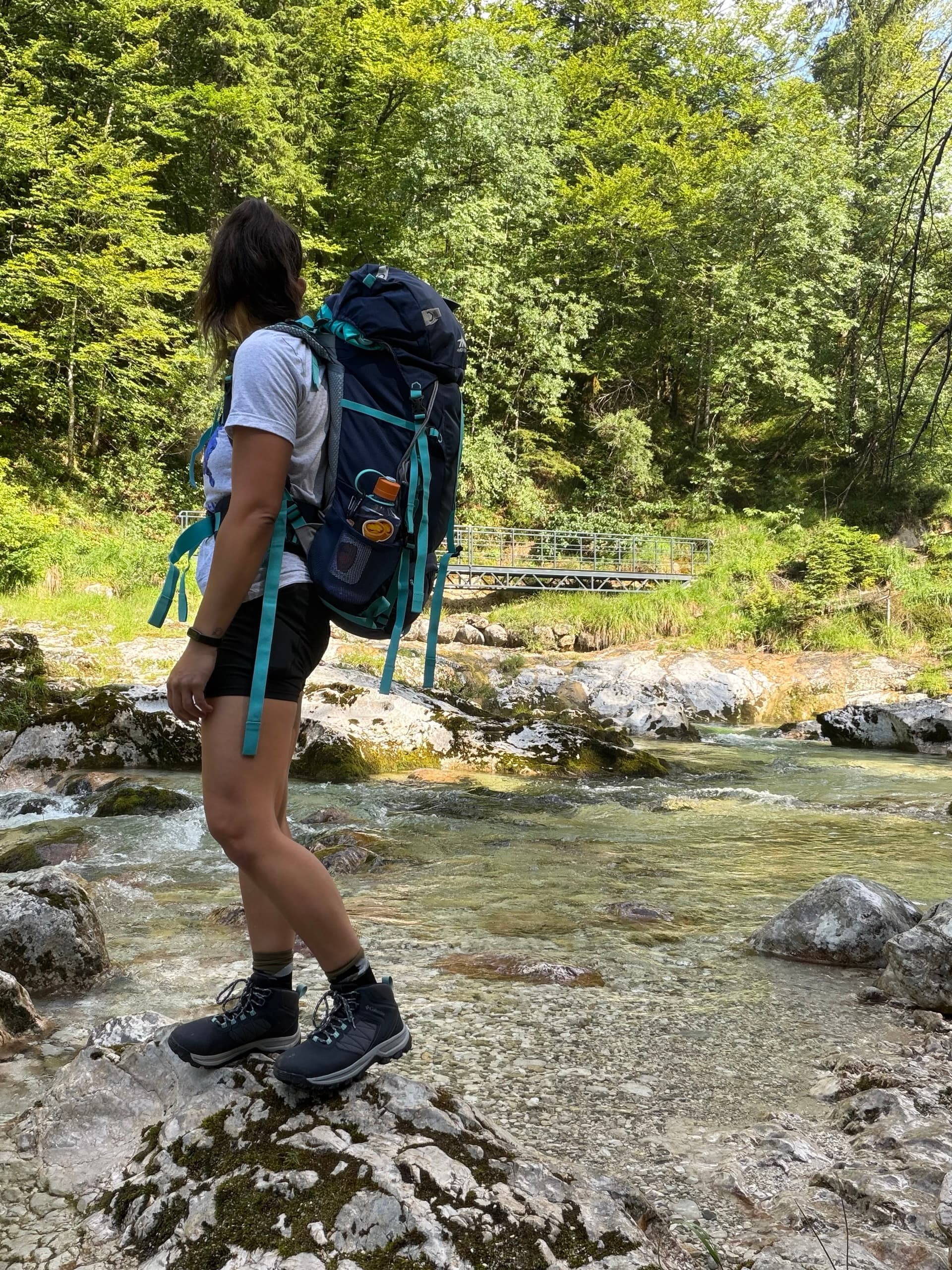 Hiker with large backpack beside clear mountain stream with rocky riverbed and forest background