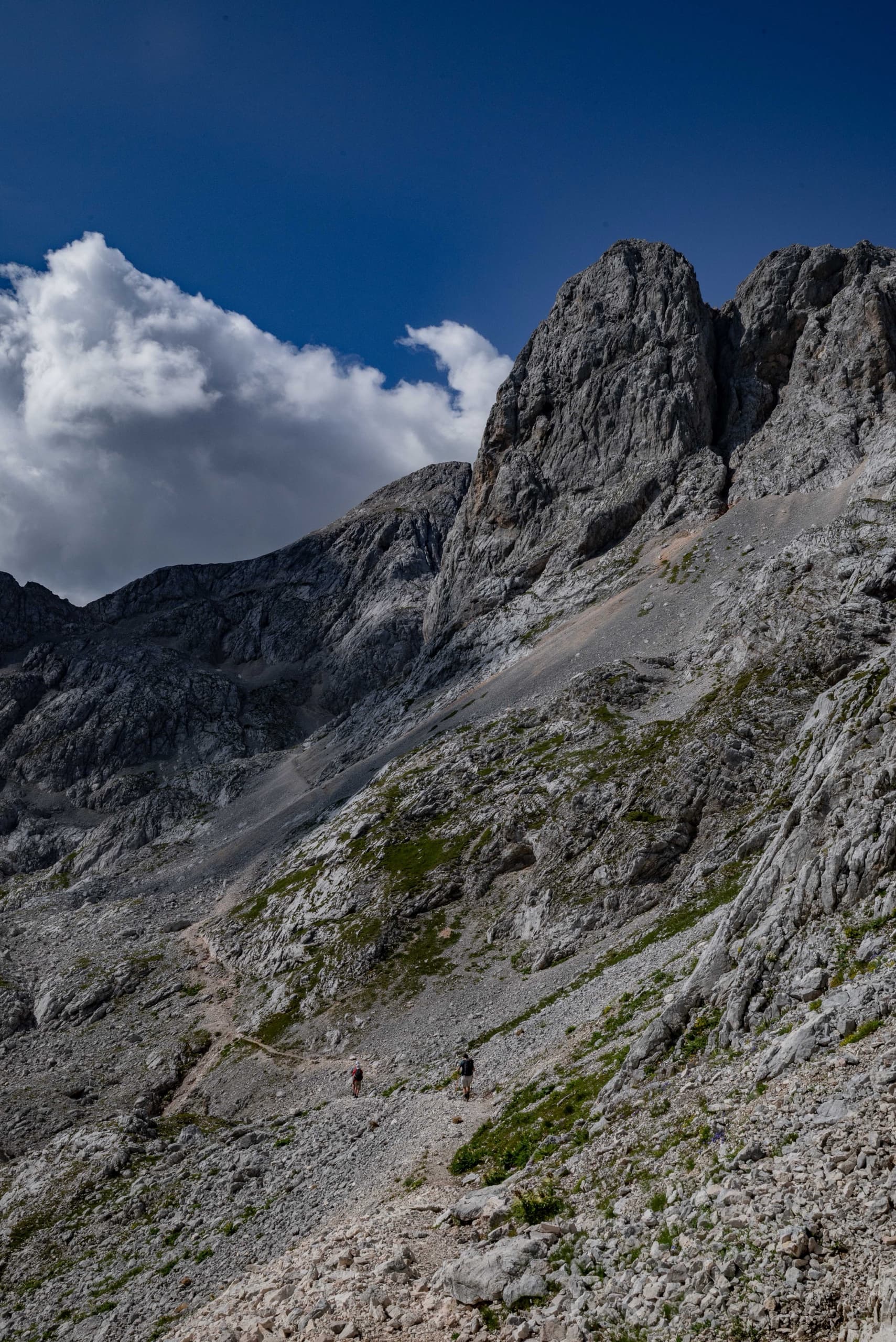 Hikers on rocky mountain trail below towering rock peaks and blue sky with clouds