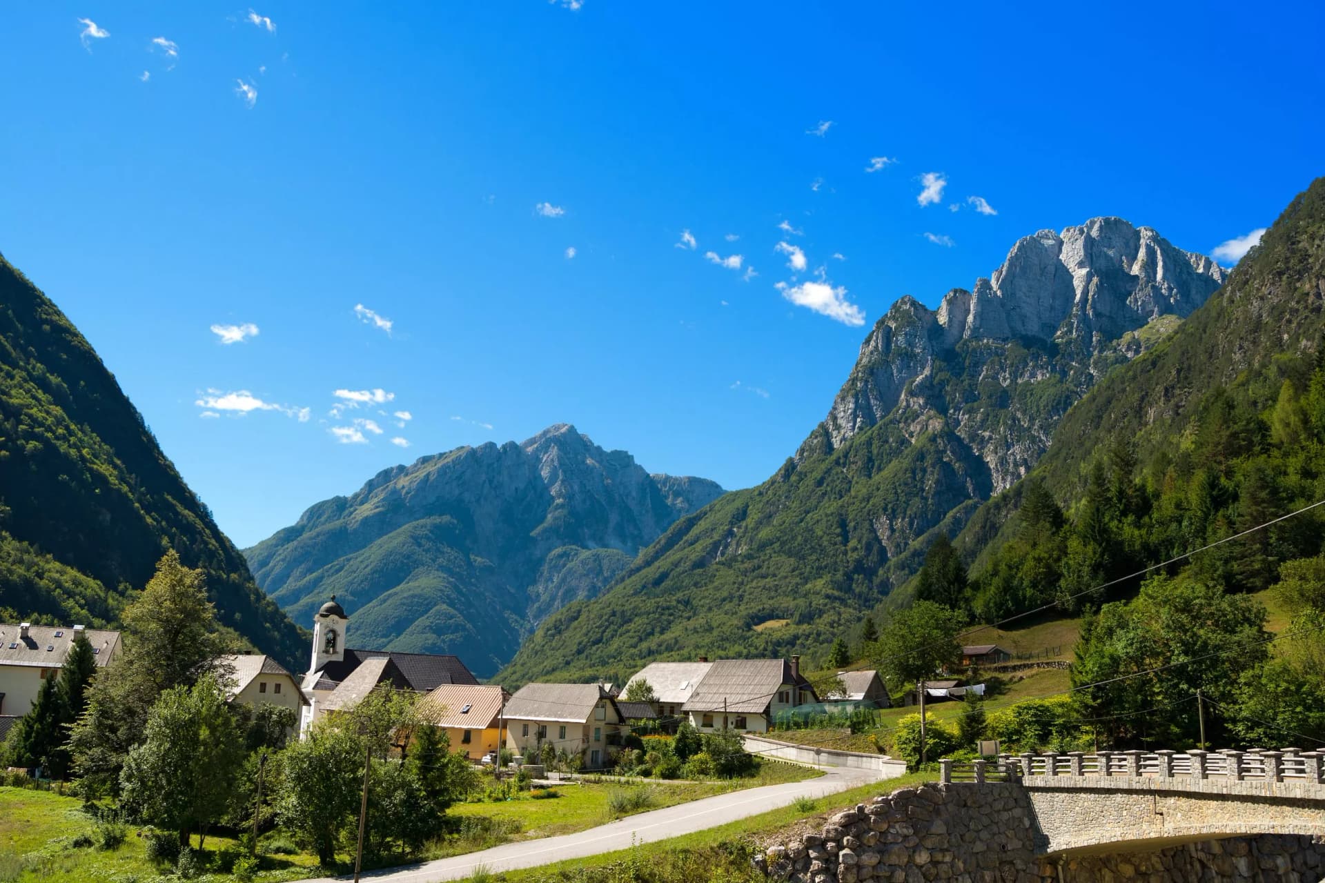 Alpine village with houses and church nestled between steep, forested mountains under a bright blue sky.